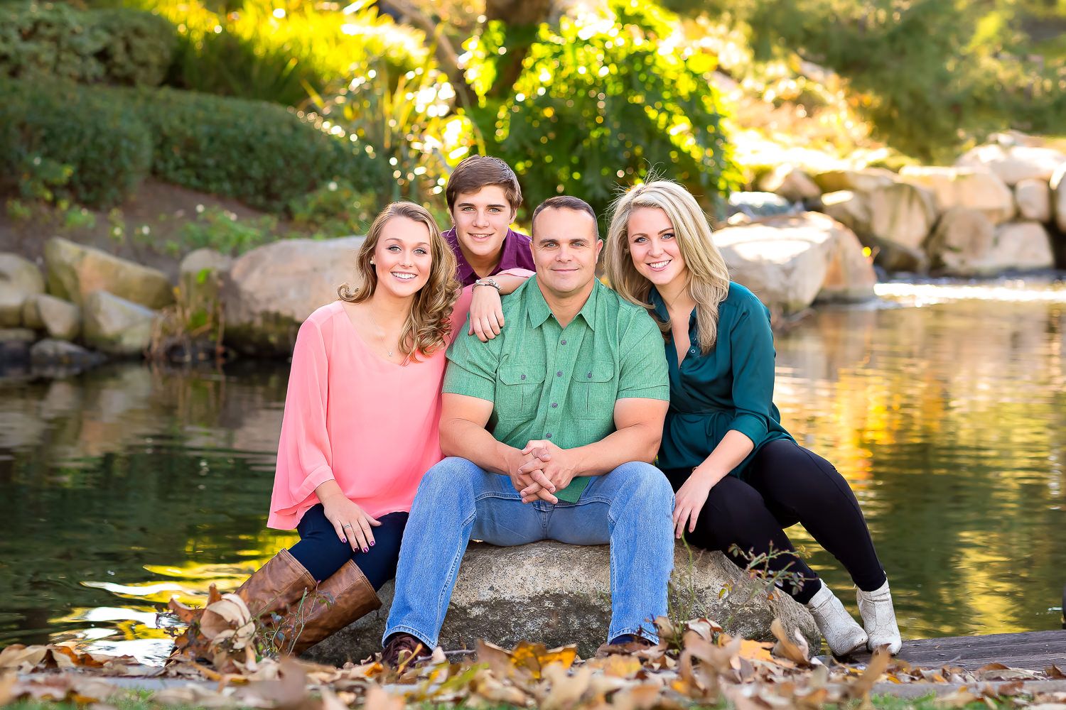 Happy family of four sitting on rocks near a pond surrounded by autumn foliage in outdoor portrait.