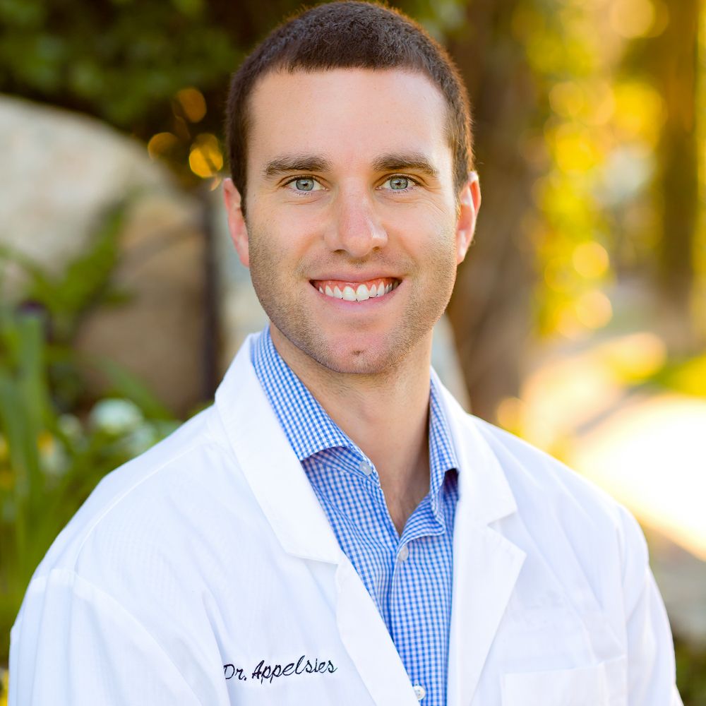 A smiling male doctor in a white lab coat with name tag, wearing a blue shirt, posing outdoors in natural light.