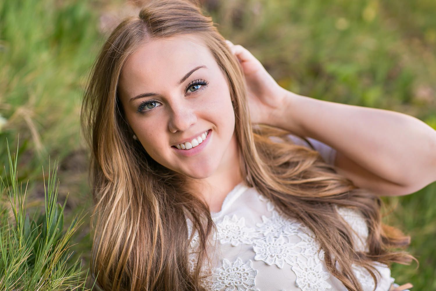 Smiling young woman with long brown hair wearing a white lace top, posing outdoors in a green field.