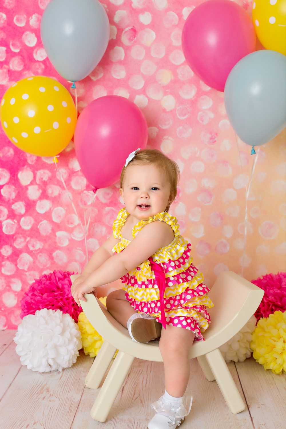 Cheerful baby girl in yellow ruffled dress sitting among colorful balloons on pink backdrop.