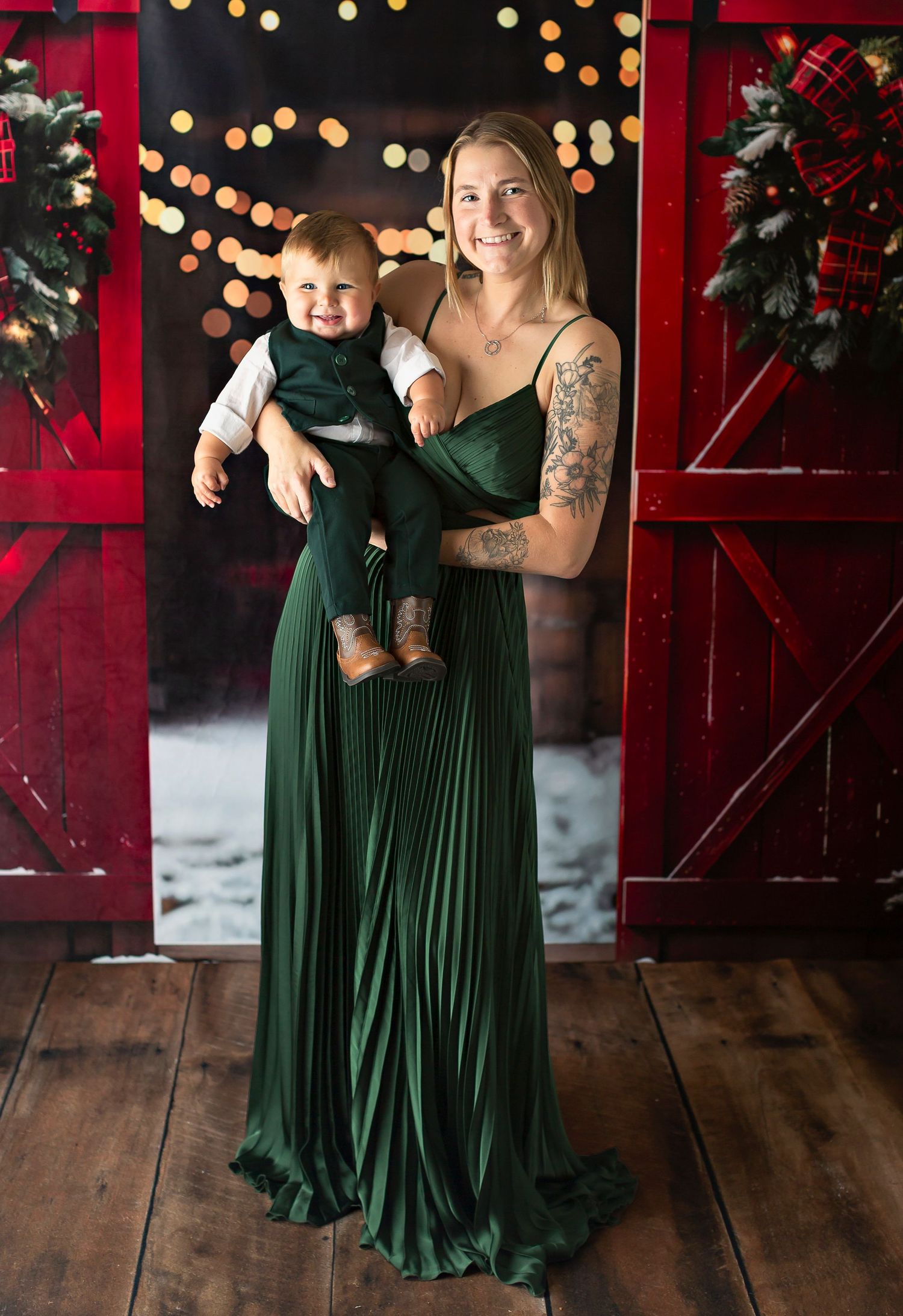 Woman in green gown holds smiling baby in matching outfit before festive Christmas backdrop with bokeh lights.