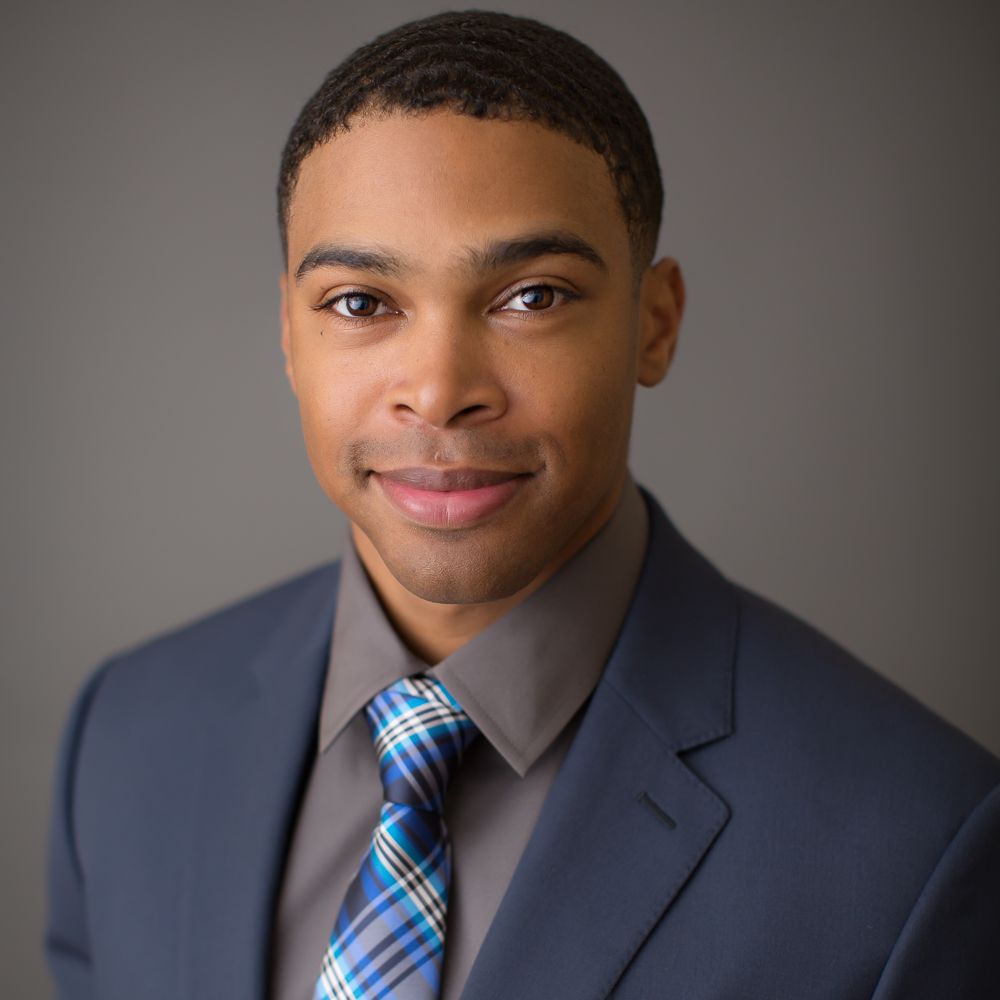 Professional headshot of a young man in a blue suit and plaid tie, smiling confidently against a gray background.