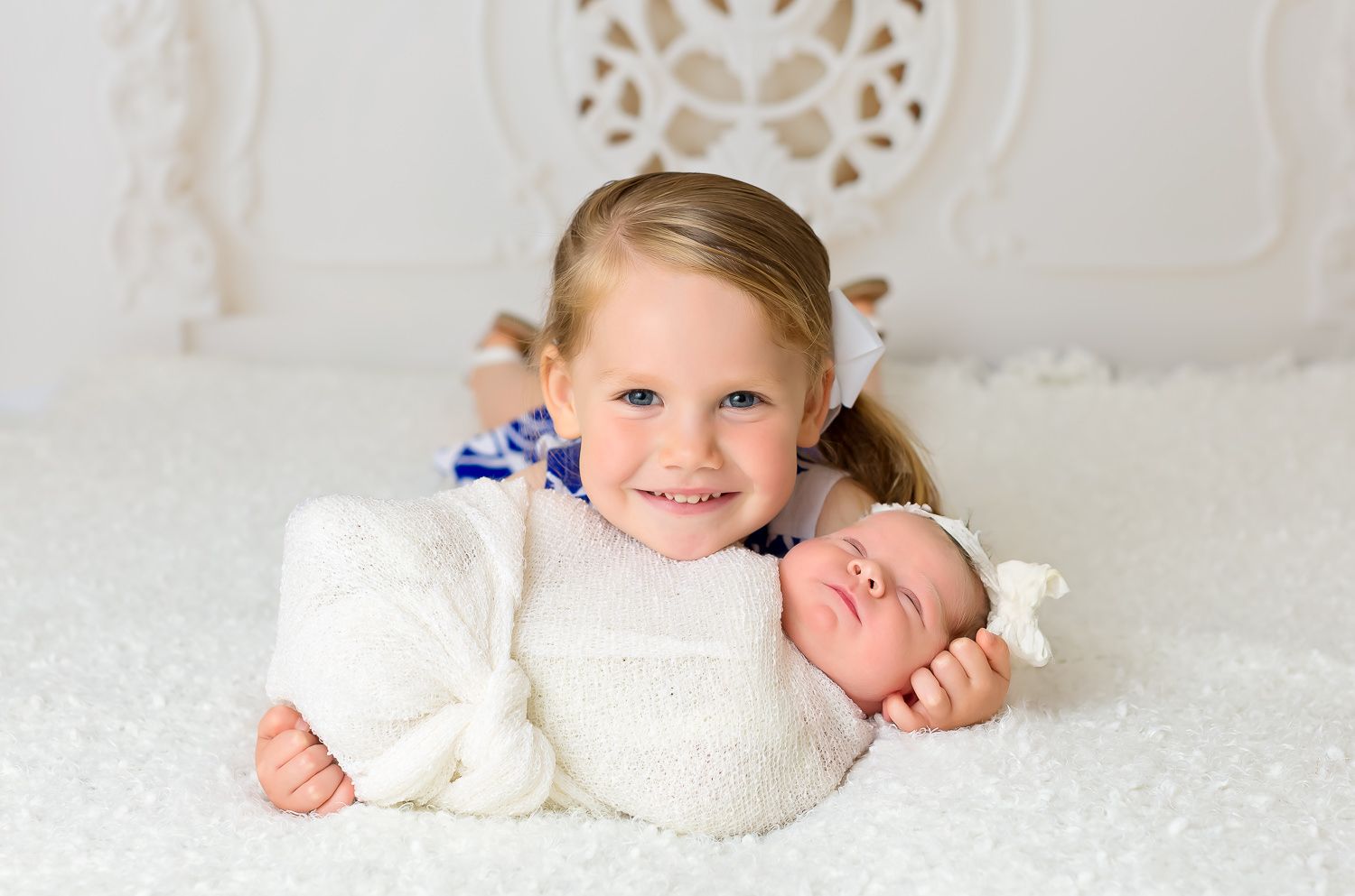 Smiling toddler girl in white dress poses with sleeping newborn sibling on a fluffy white blanket.
