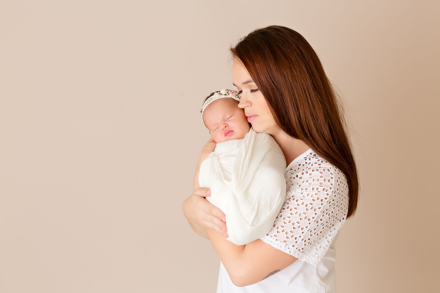 Brunette mother gently holds sleeping newborn wrapped in white against her cheek.