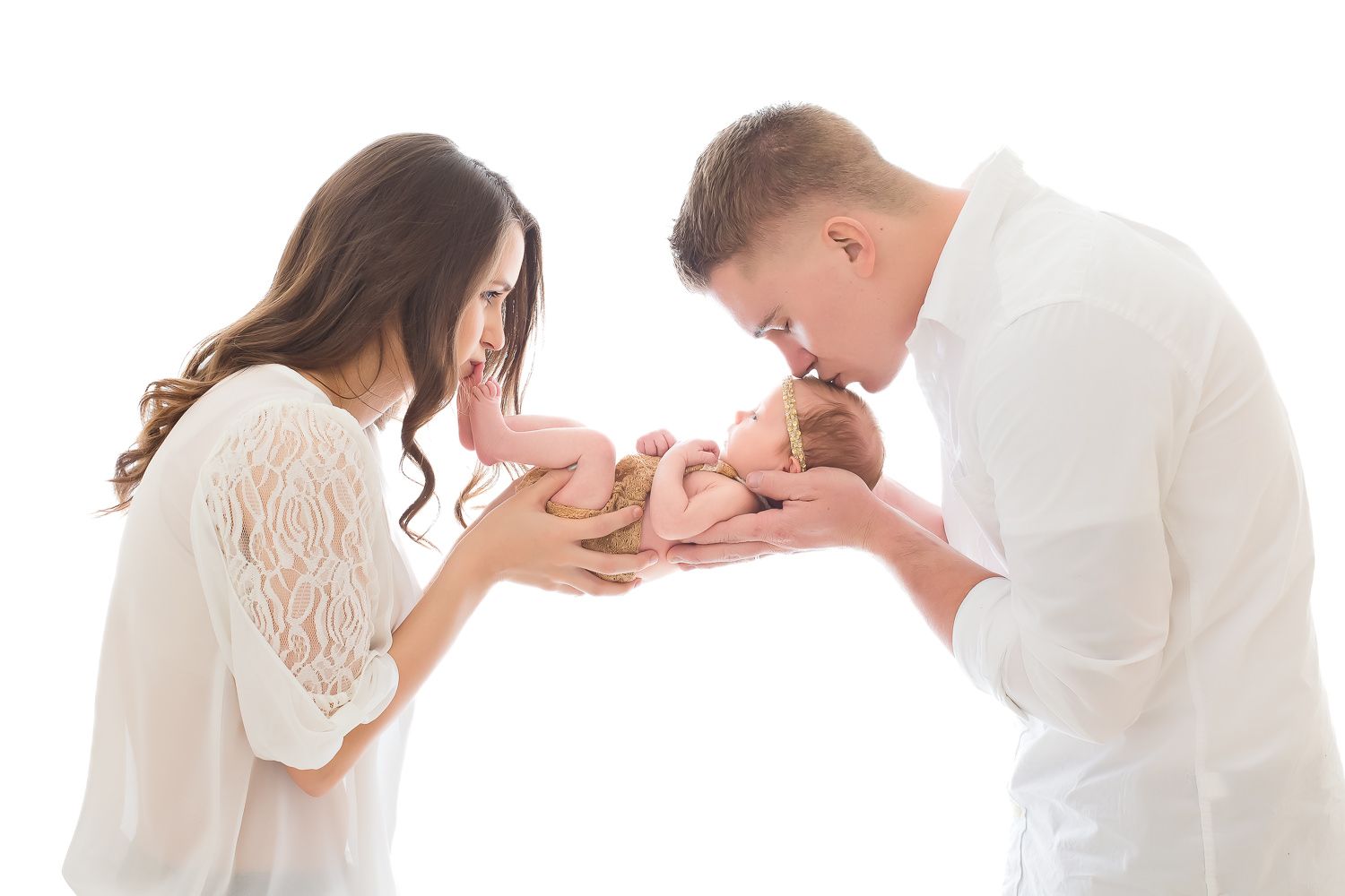 Parents in white outfits tenderly kissing their newborn baby against white background.