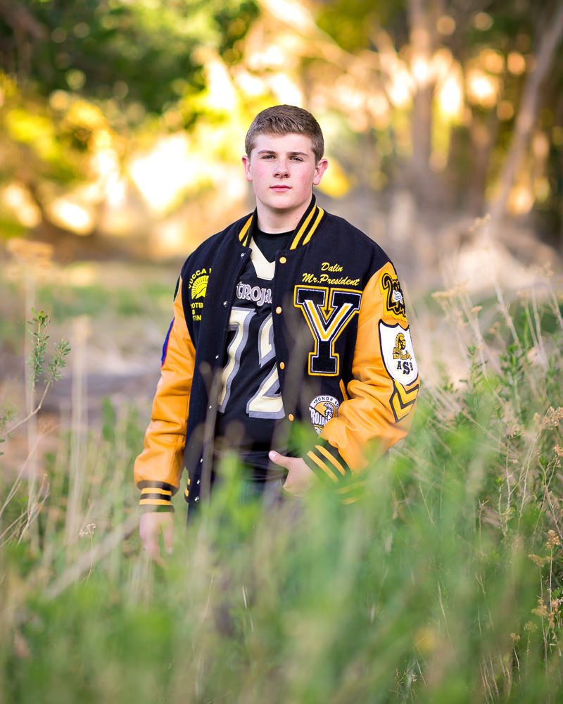 Young man in black and gold varsity letterman jacket standing in tall grass with trees in background.