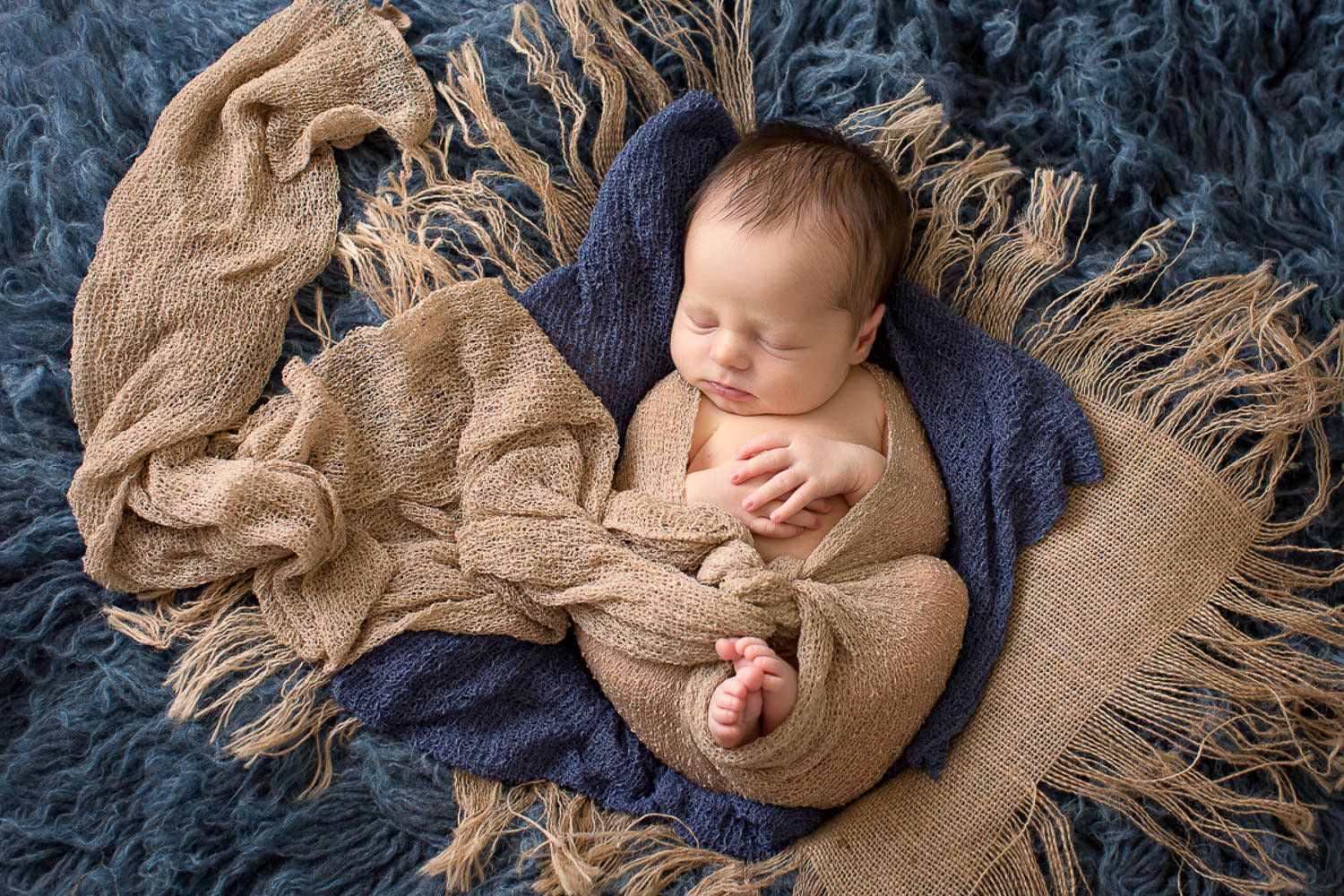 Sleeping newborn baby wrapped in blue and tan blankets in rustic bowl.