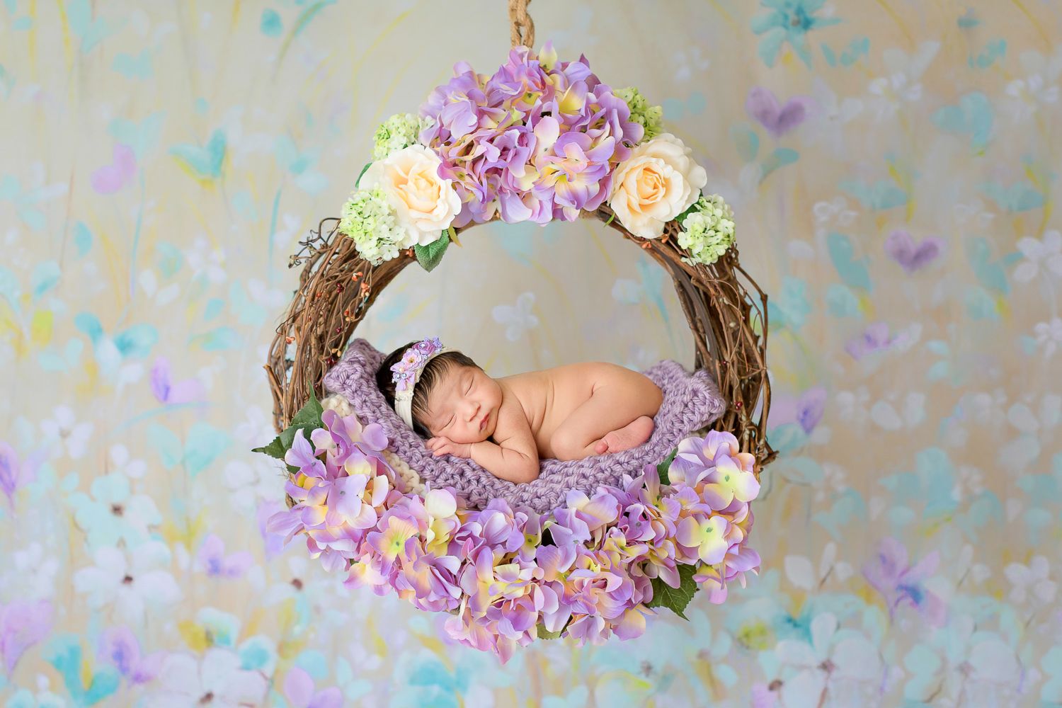 Newborn baby sleeping peacefully in a floral wreath swing decorated with purple hydrangeas and roses.