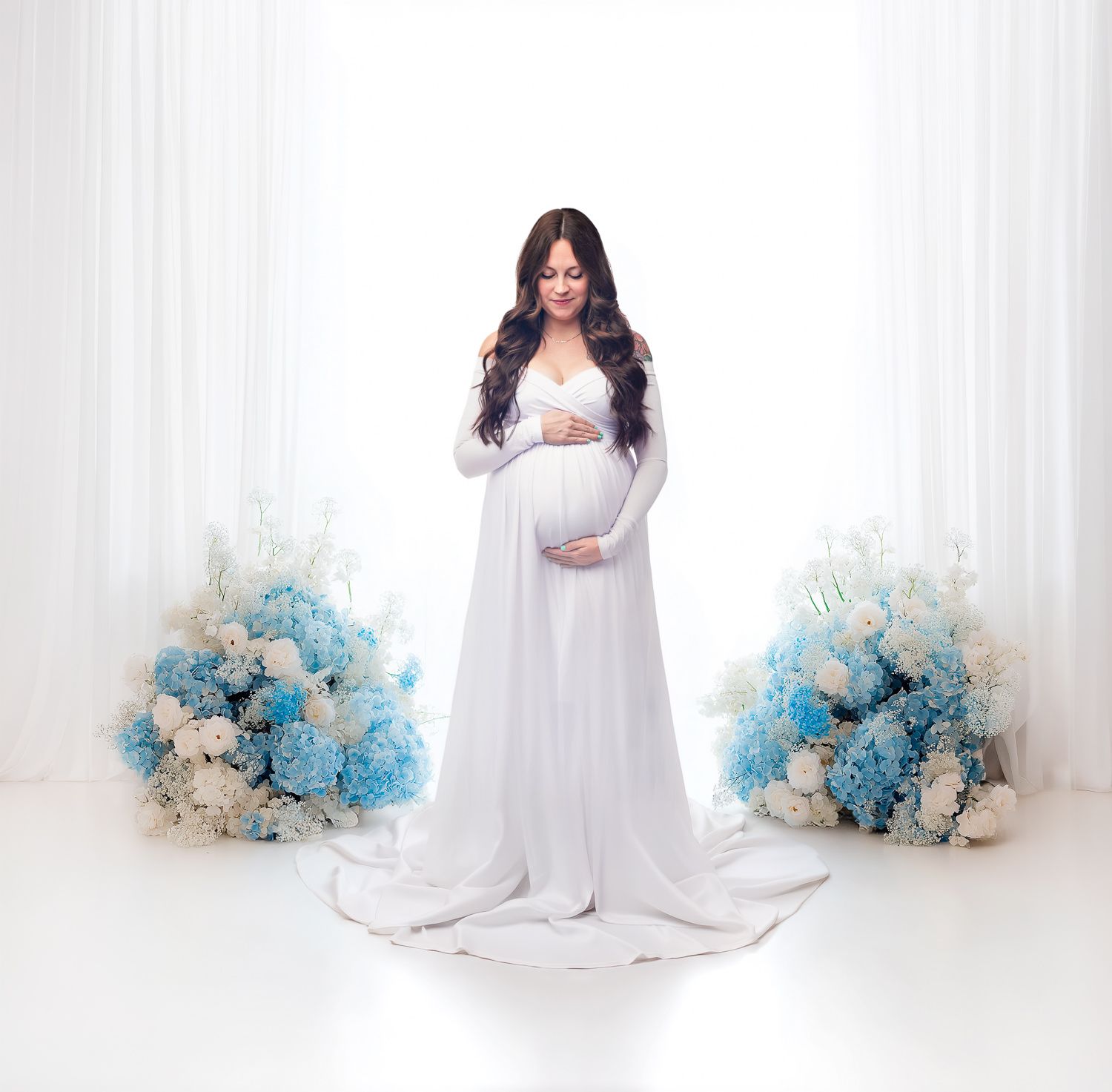 Pregnant woman in white flowing gown surrounded by blue and white flowers.