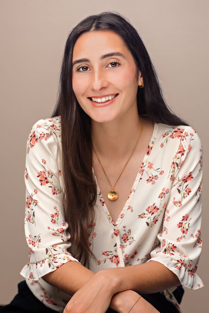 Smiling young woman with long dark hair wearing a floral blouse and gold locket necklace, posed against a beige background.