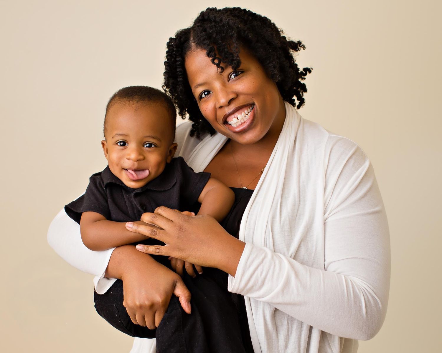 Smiling Black mother holding her happy baby boy, both dressed smartly, against a neutral beige background.