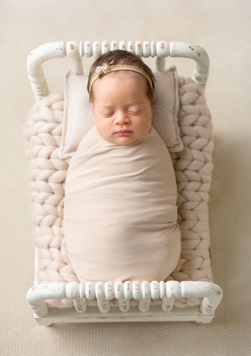Peacefully sleeping newborn baby swaddled in soft blush wrap, wearing gold floral headband, lying on miniature white crib.