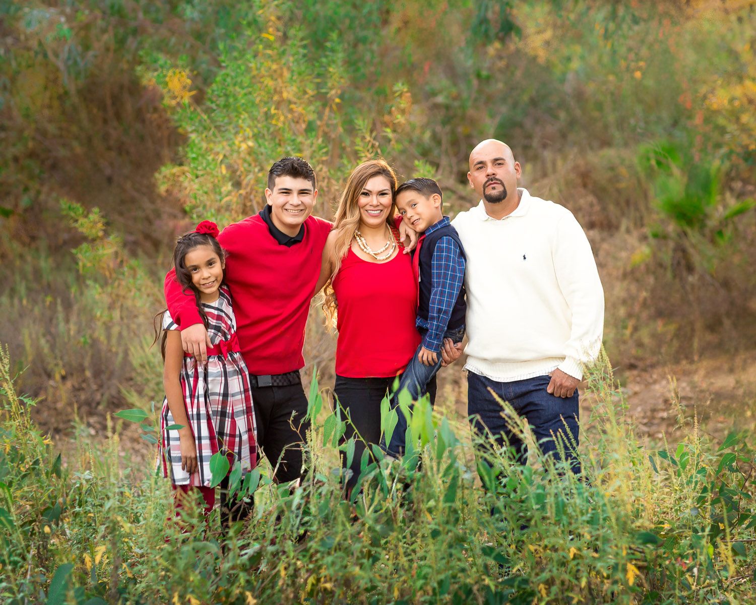 Family of five posing outdoors in red and white outfits surrounded by lush green foliage for a holiday portrait.