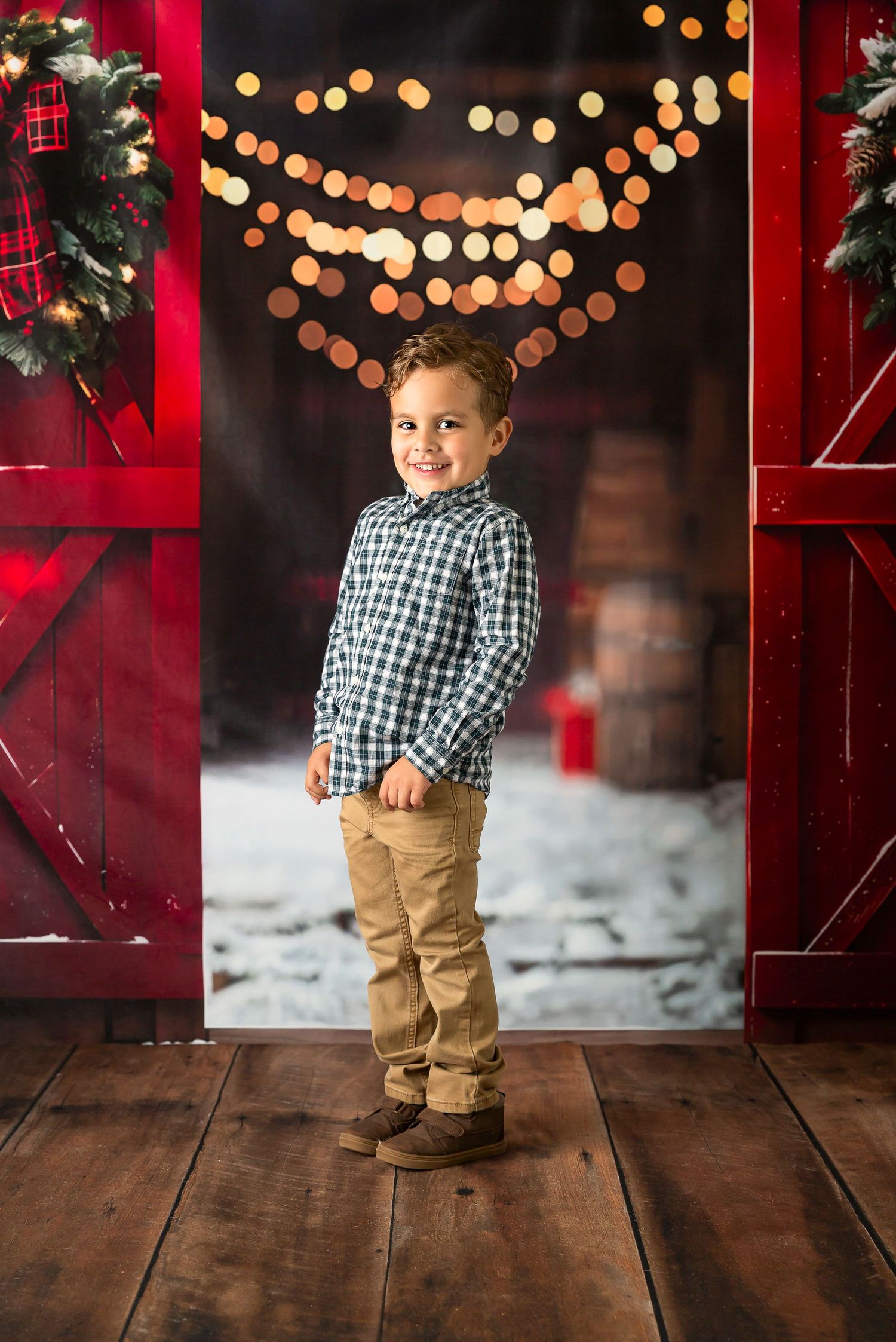 A smiling young boy in a plaid shirt and khaki pants poses in a festive red barn doorway with bokeh Christmas lights.