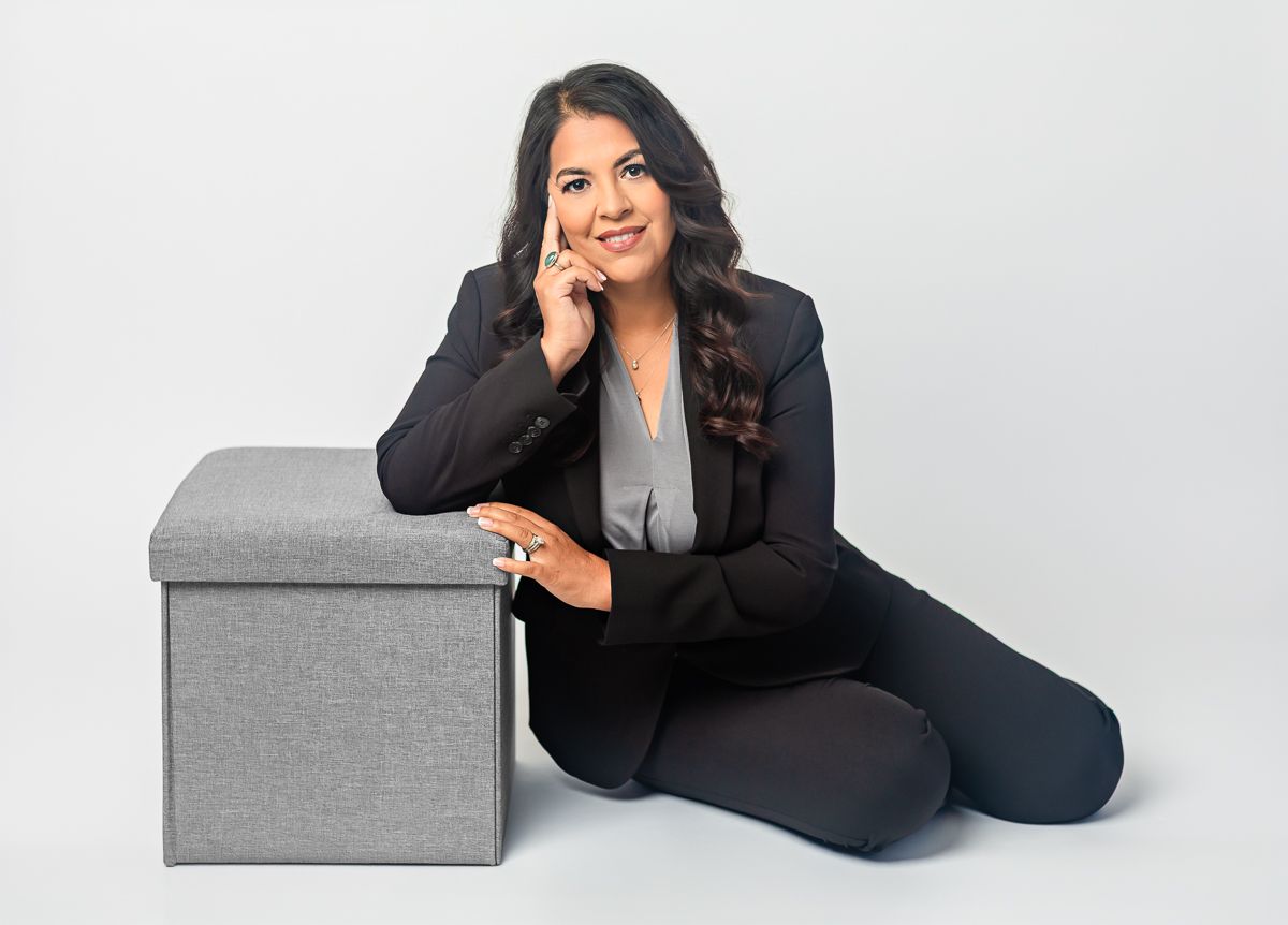 Professional woman in black blazer sitting beside a gray storage ottoman, posing confidently against white background.