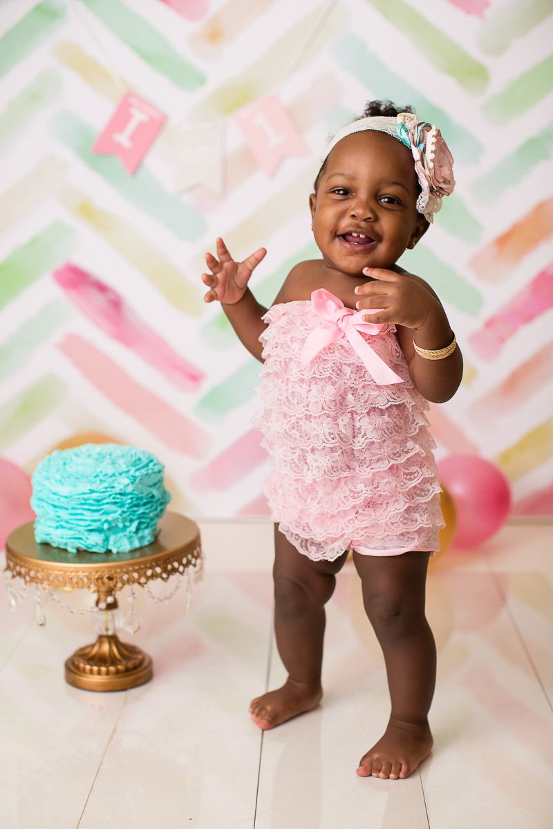 Joyful baby girl in pink lace romper standing beside teal smash cake on gold stand.