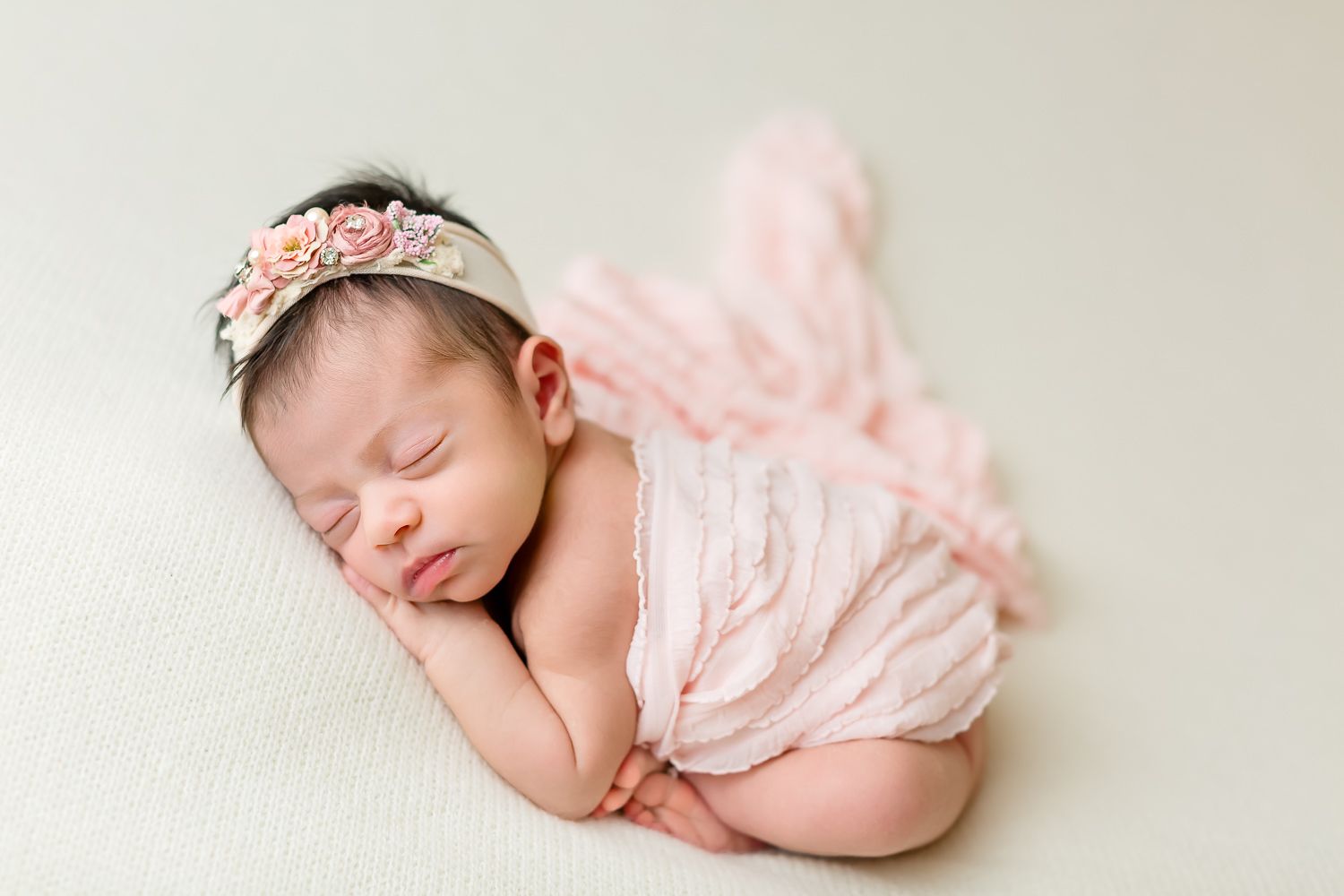 Sleeping newborn baby girl in pink wrap and floral headband posed on cream surface.