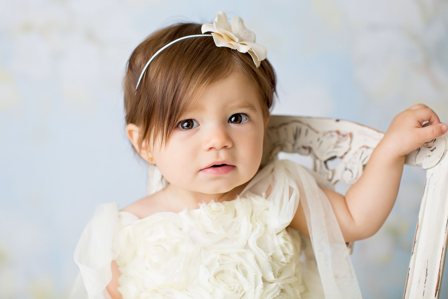 Sweet baby girl in white floral dress with headband posing for professional studio portrait.