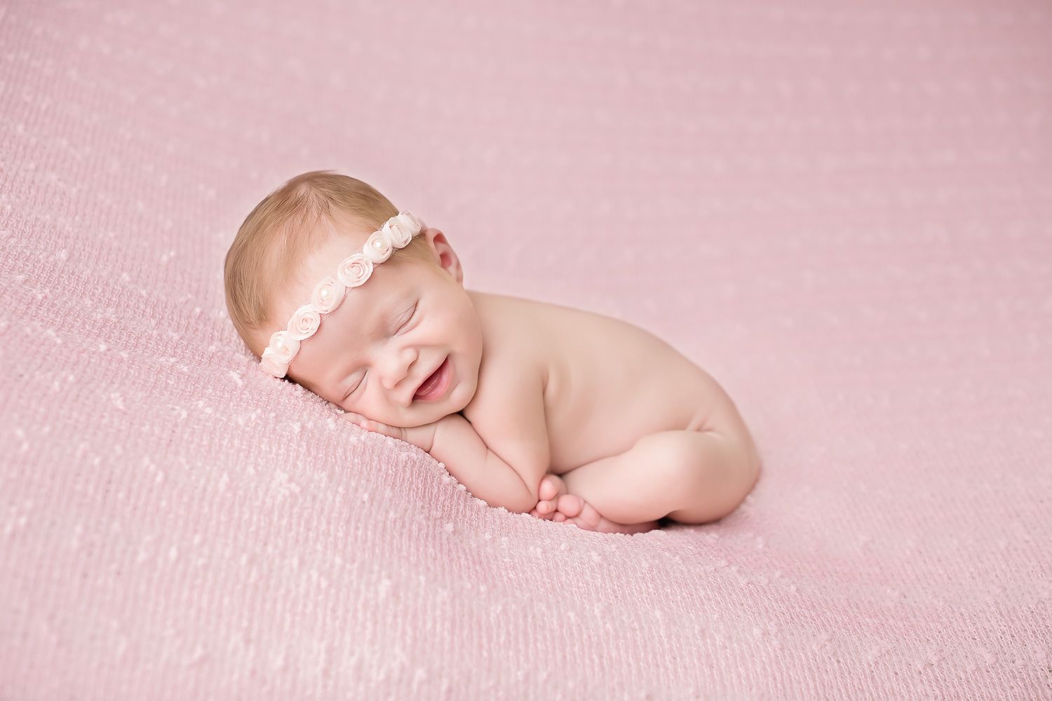 Sleeping newborn baby girl in pink headband on pink blanket smiling.