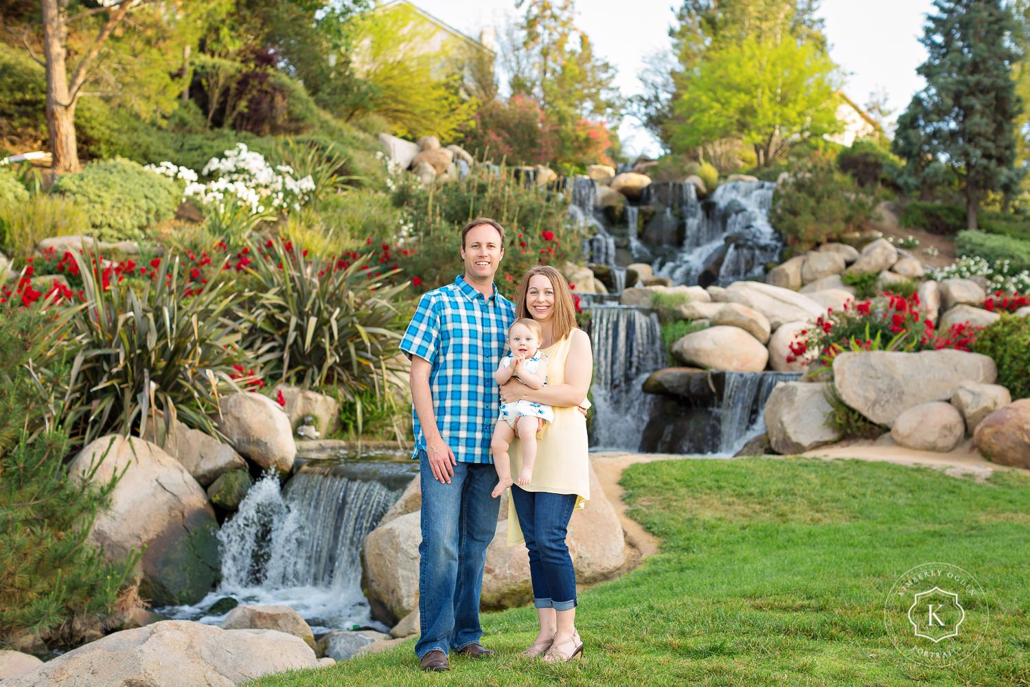 Smiling couple holding baby standing in front of beautiful waterfall garden landscape in outdoor family portrait.