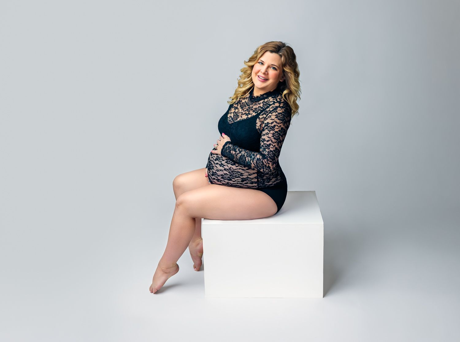 Pregnant woman in black lace bodysuit sitting on white cube in studio.