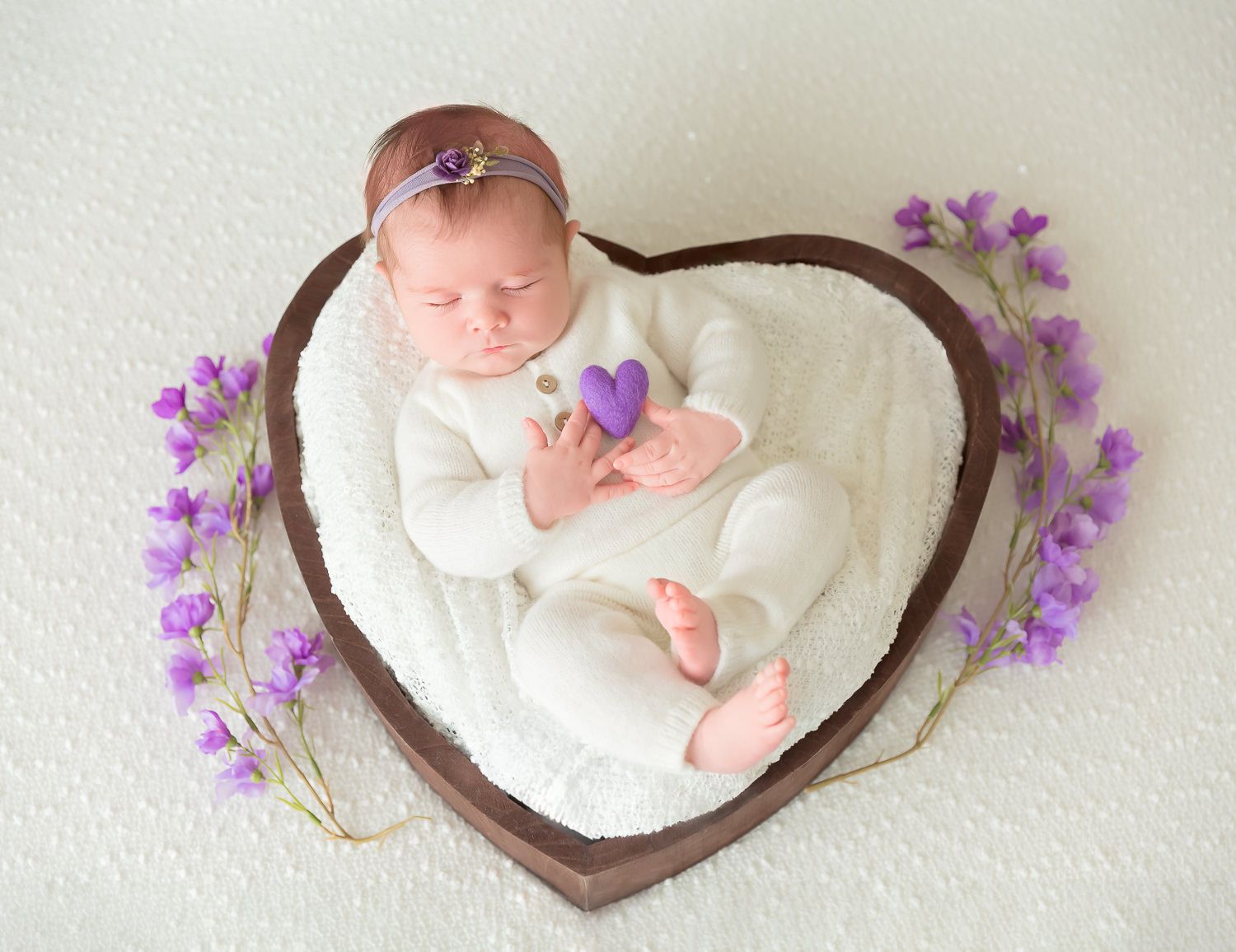 Newborn baby in white outfit sleeps in heart-shaped wooden bowl surrounded by purple wildflowers.