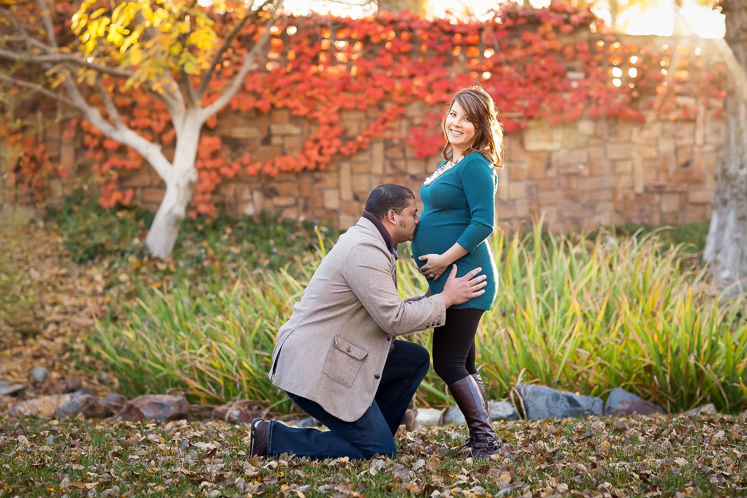 Man kneeling to embrace pregnant woman's belly outdoors in autumn garden with colorful foliage.