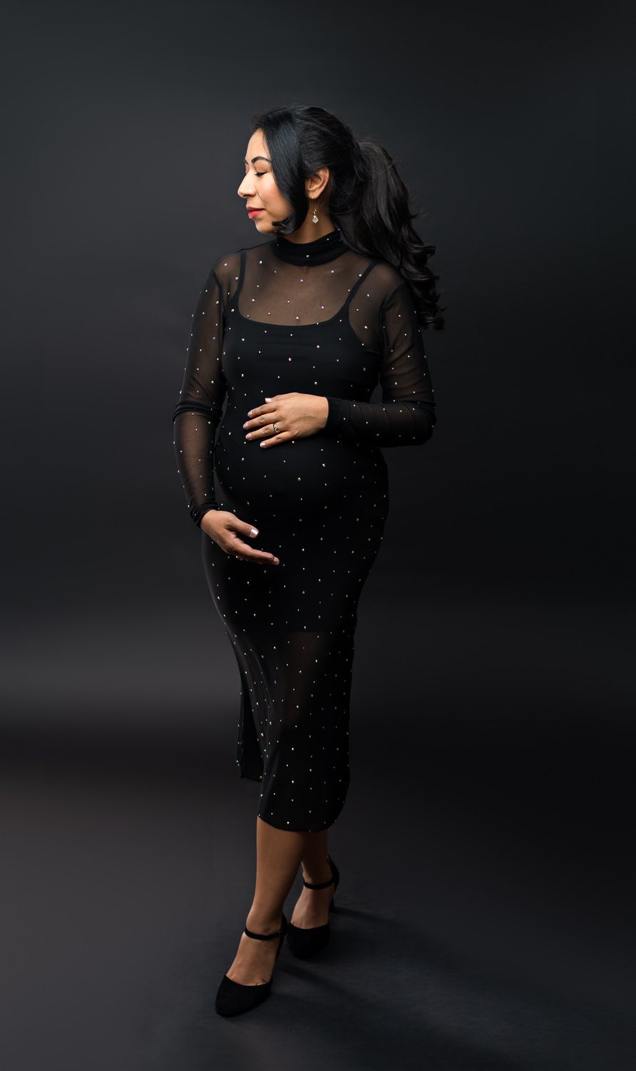 Pregnant woman in elegant black mesh midi dress with sparkles, posing in studio against dark background.