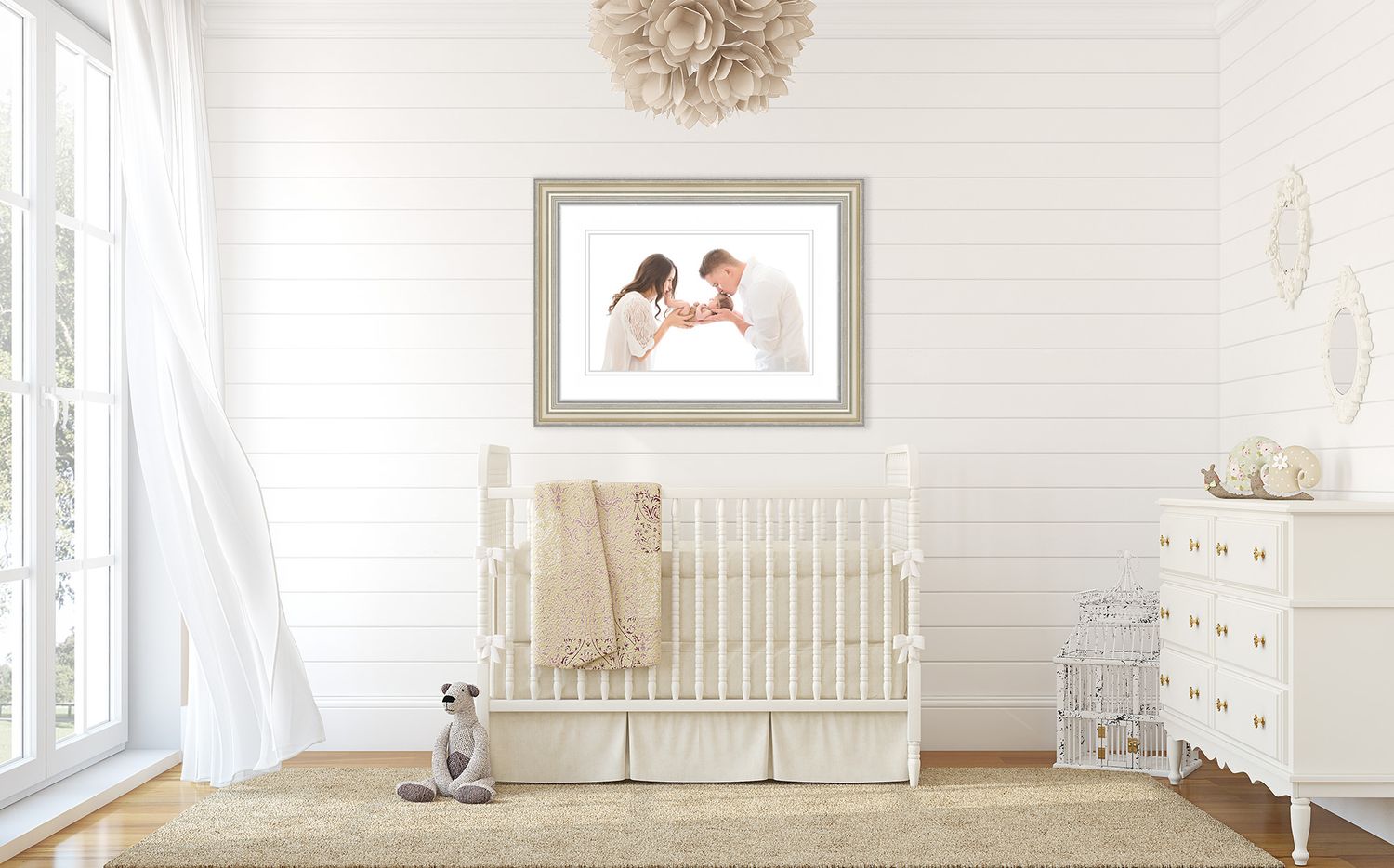 Framed newborn family portrait displayed above white crib in elegantly decorated nursery room.