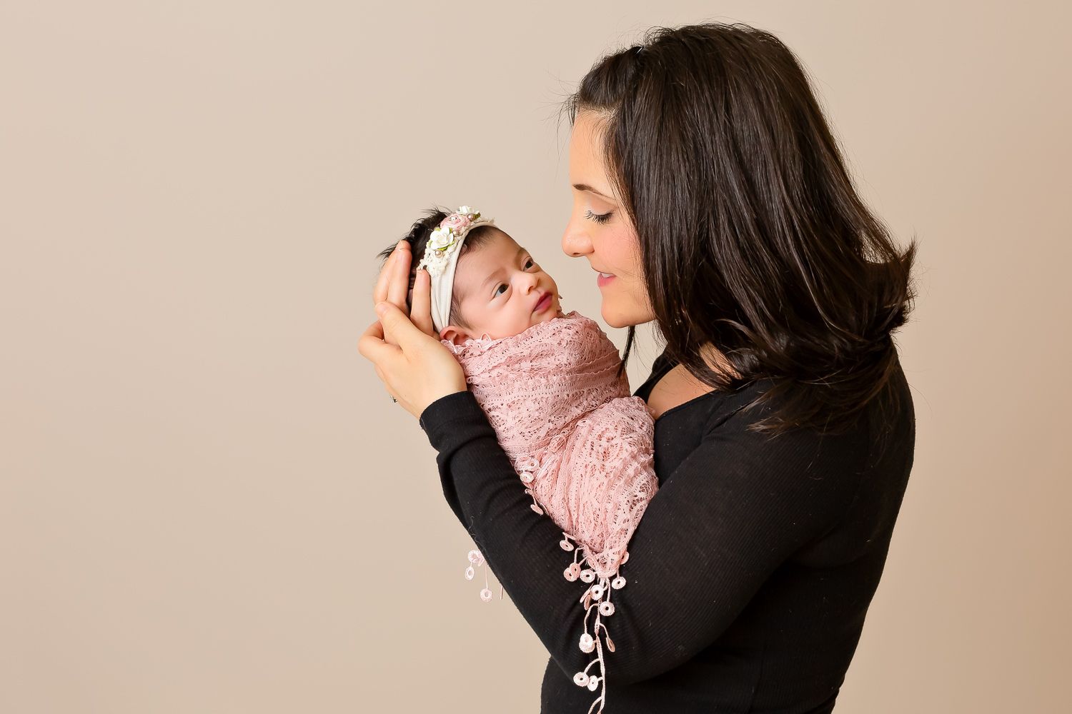 Loving mother in black top holding up smiling newborn baby wrapped in pink floral swaddle.