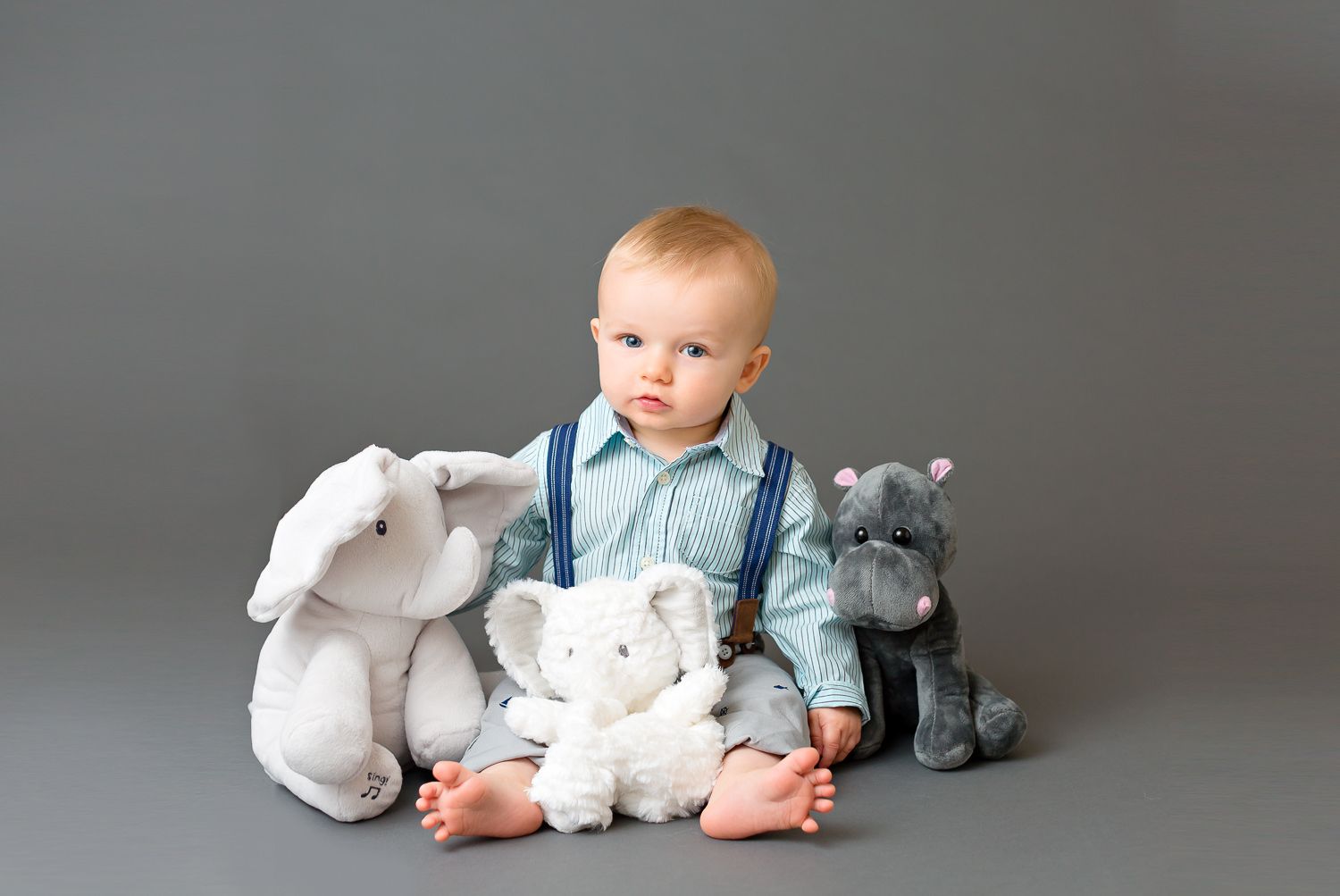 Baby boy in suspenders sitting with three stuffed animal toys on gray background.