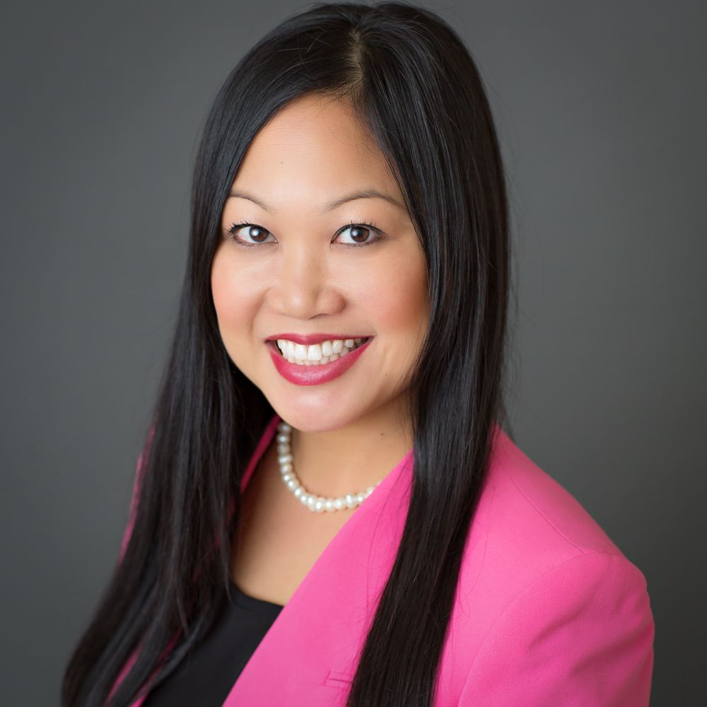 Professional headshot of a smiling woman in a pink blazer and pearl necklace against a gray background.