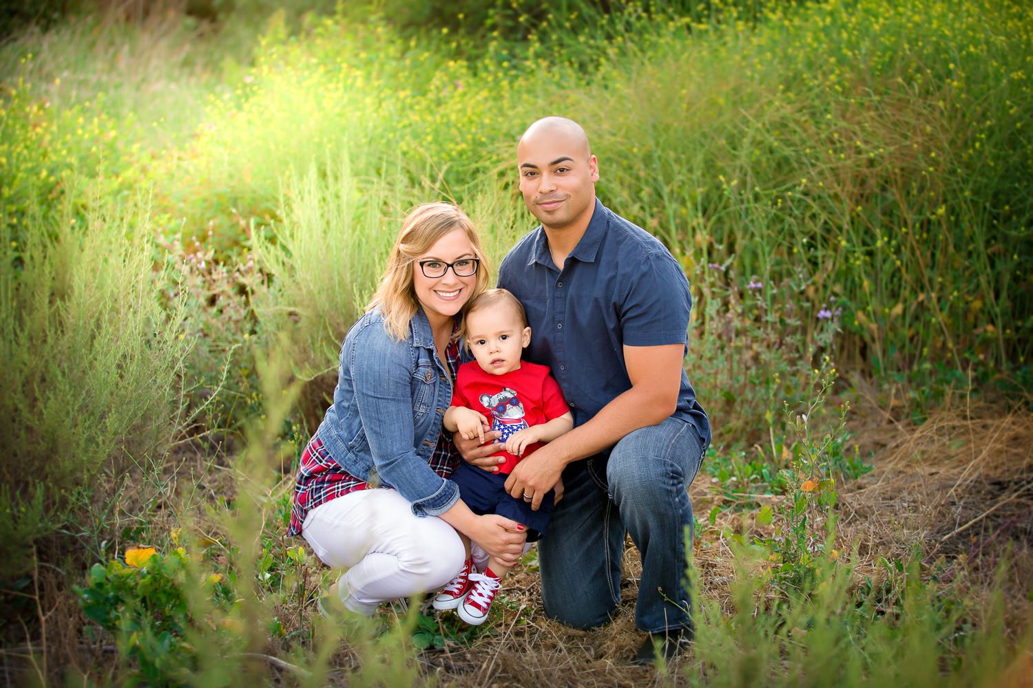 Young family with baby posing together in golden sunlit meadow field for outdoor nature portrait session.