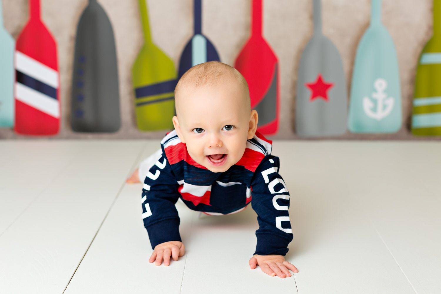 Smiling baby boy in navy nautical outfit crawling on white floor with colorful oar backdrop.