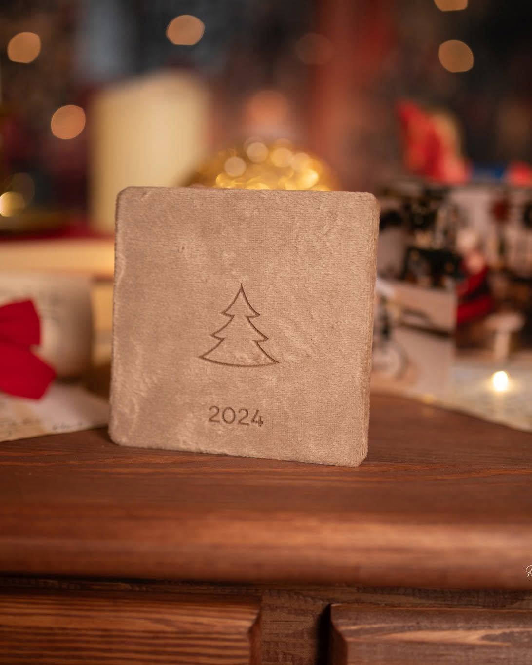 A square stone coaster engraved with a Christmas tree and 2024, displayed on a wooden surface with festive holiday decor.