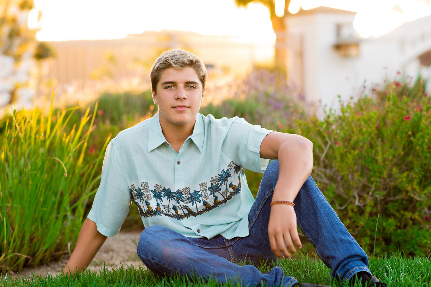 A teenage boy in a light blue Hawaiian shirt sits on green grass outdoors during golden hour.