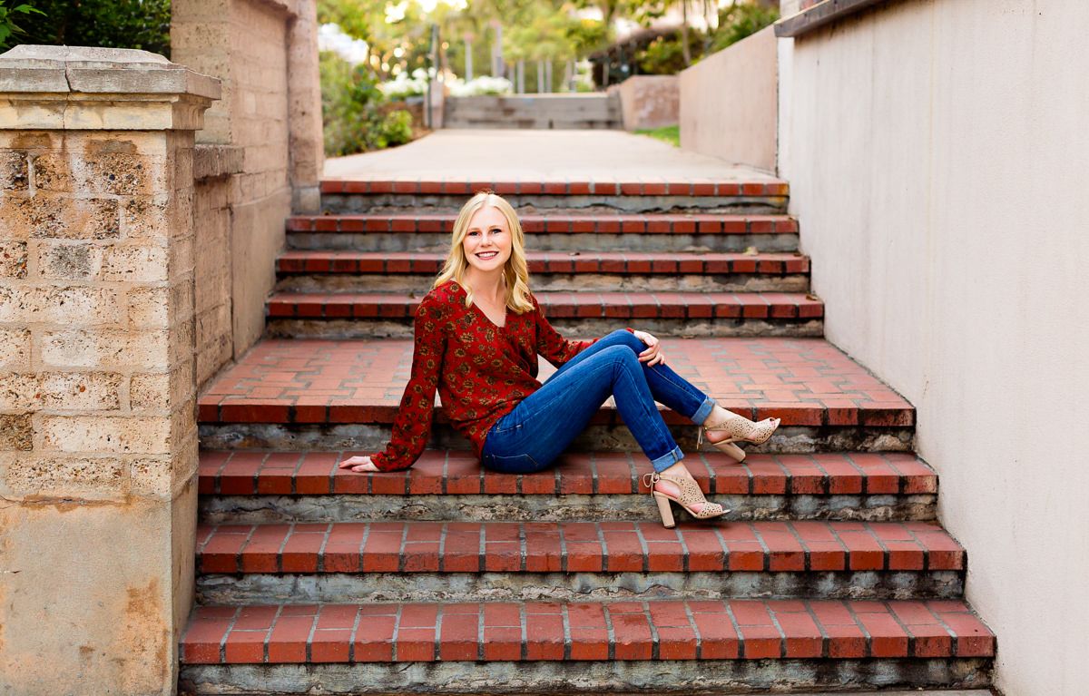 Blonde woman in red top and jeans sitting on brick stairs outdoors in a sunny, landscaped setting.