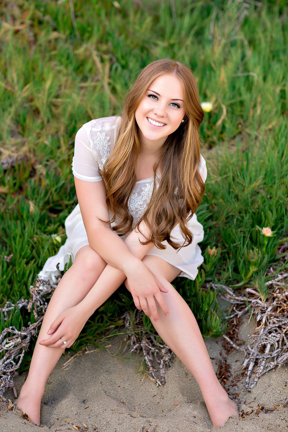 Young woman with long brown hair in white dress sitting barefoot on sandy beach with green grass backdrop.
