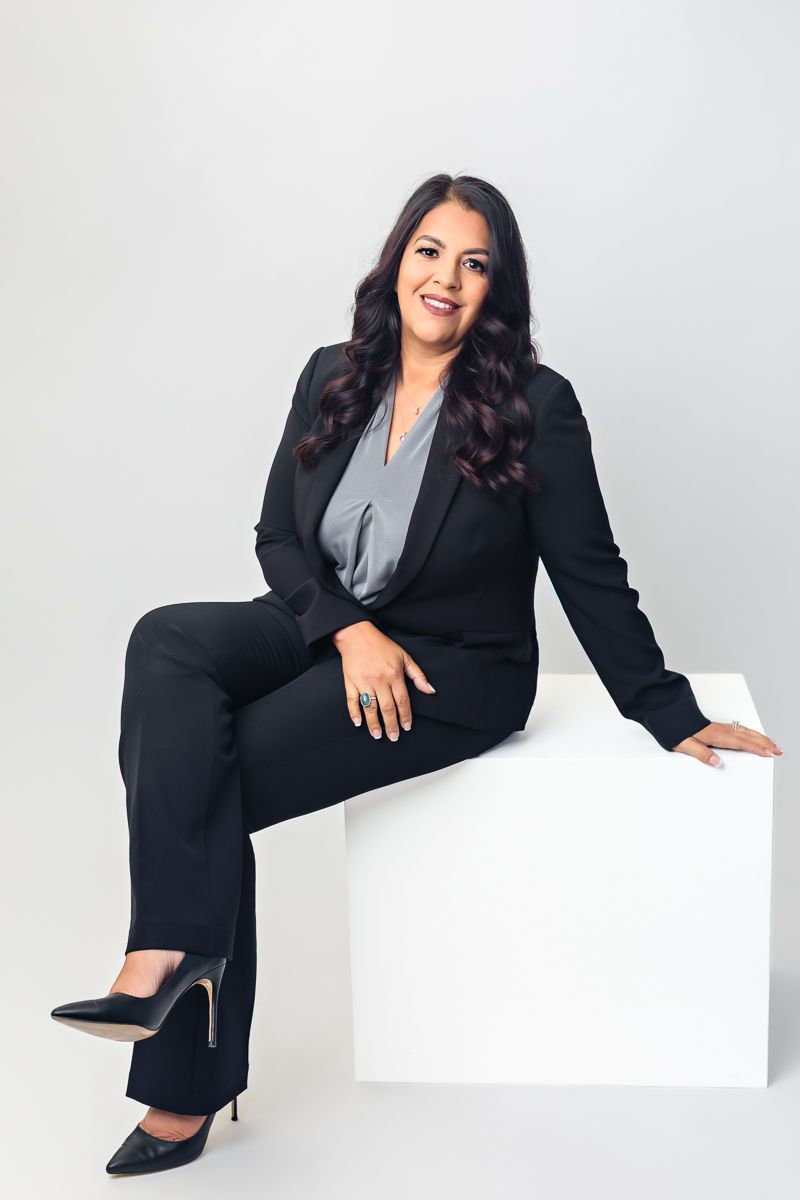 A professional woman in a black business suit sits confidently on a white cube against a light gray background.