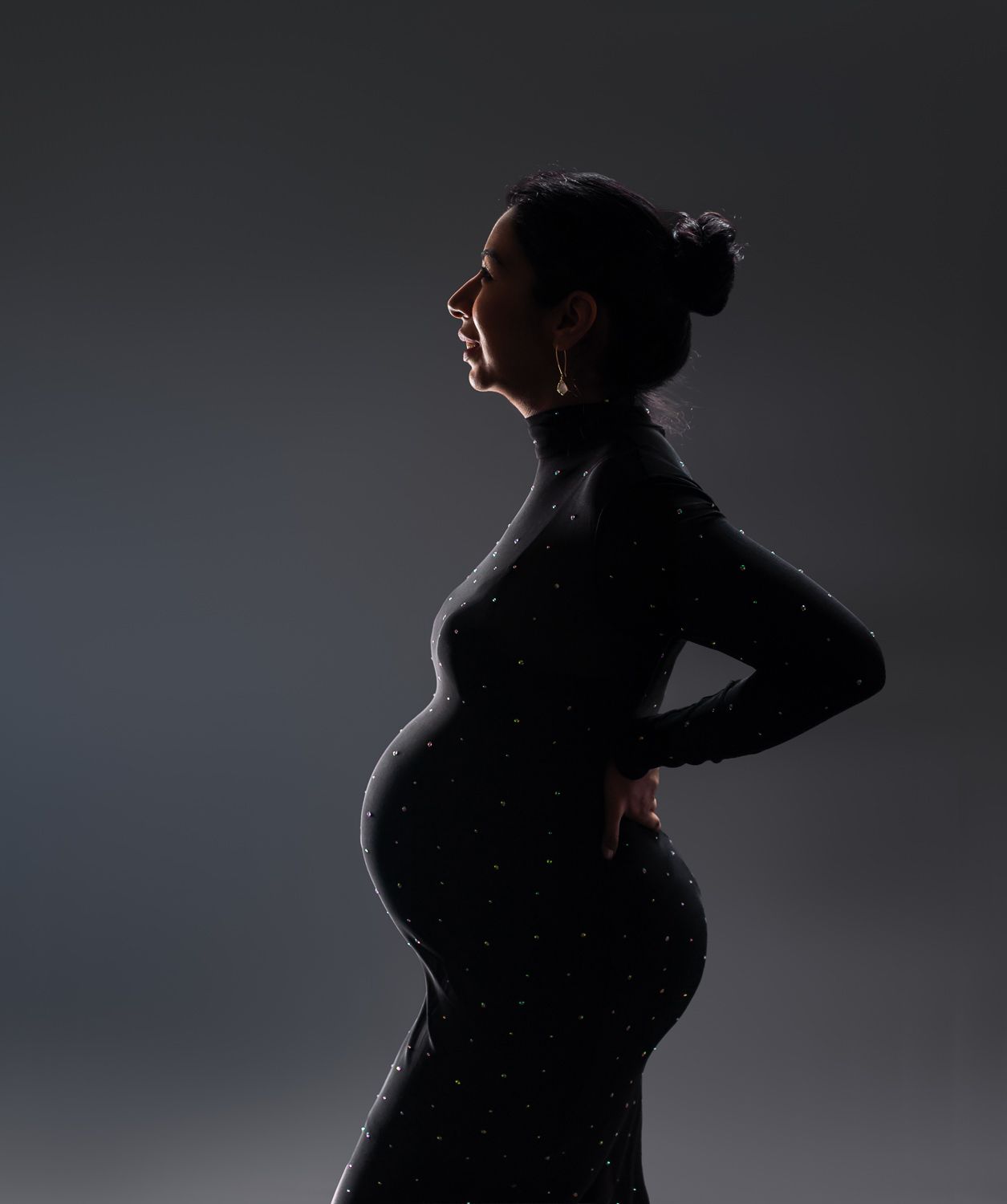 Dramatic silhouette of pregnant woman in black sparkle dress against dark background in maternity portrait.