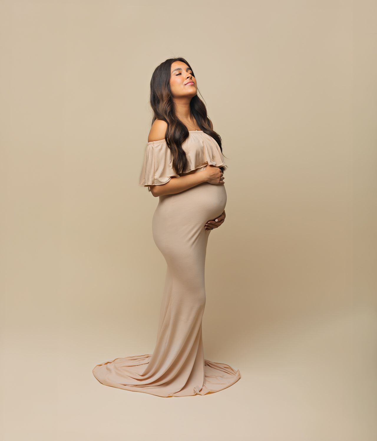 Pregnant woman in elegant beige off-shoulder maternity gown posing gracefully against neutral background.