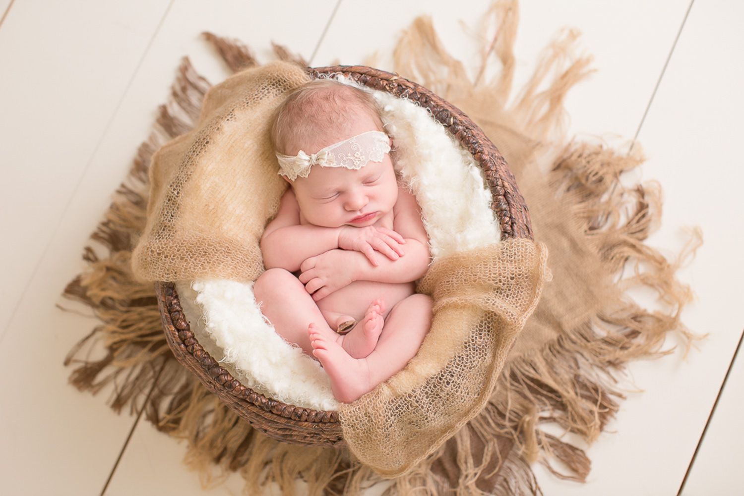 Sleeping newborn baby in pink outfit curled in rustic bowl with burlap fringe wrap.