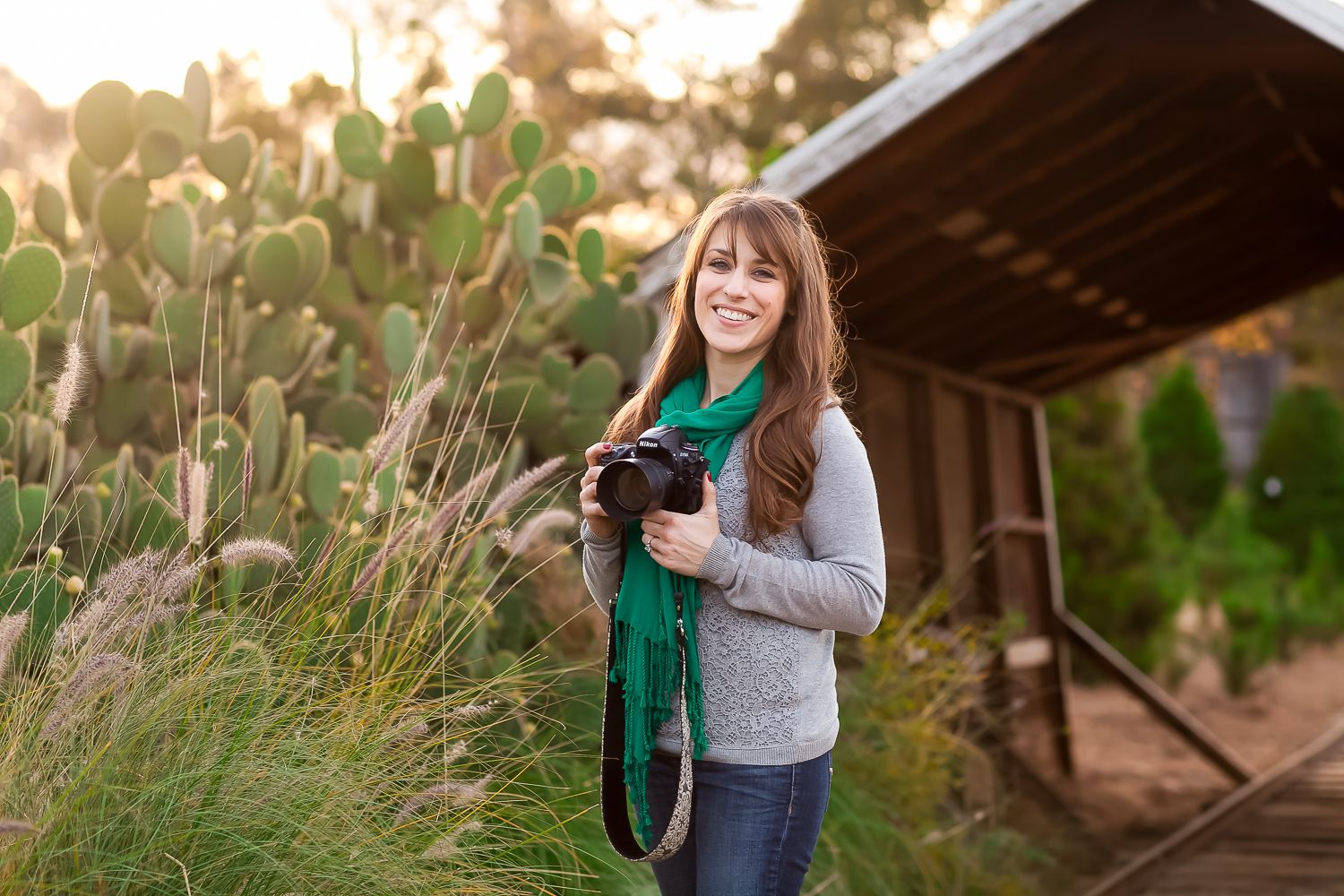 Smiling woman holding a camera with green scarf outdoors near cacti and golden sunlight.