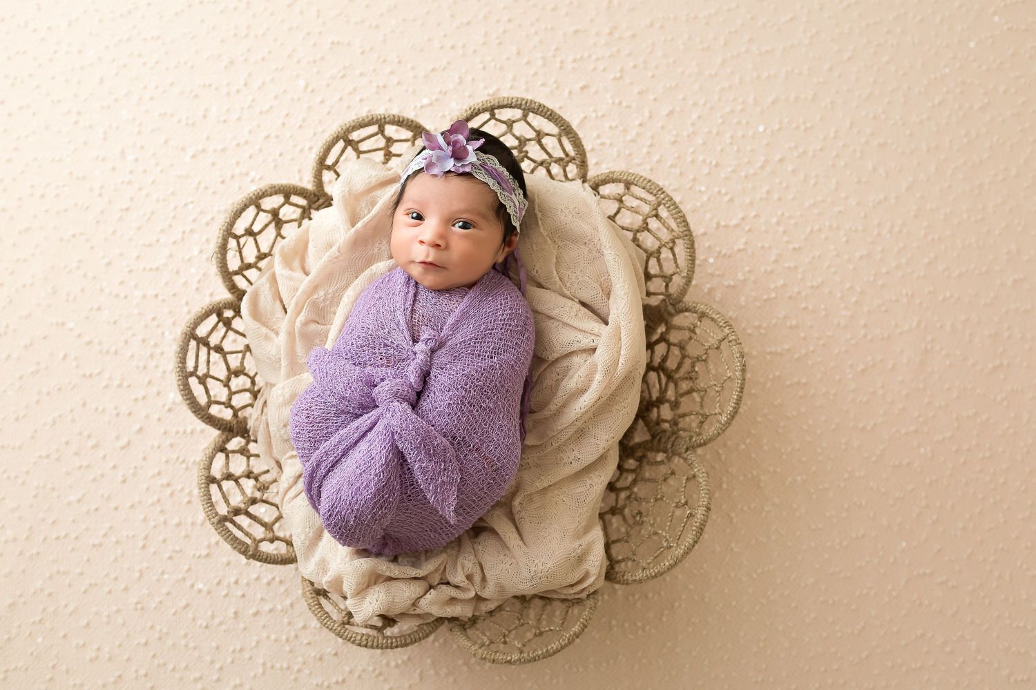 Newborn baby girl swaddled in purple wrap with bow headband in decorative floral basket.