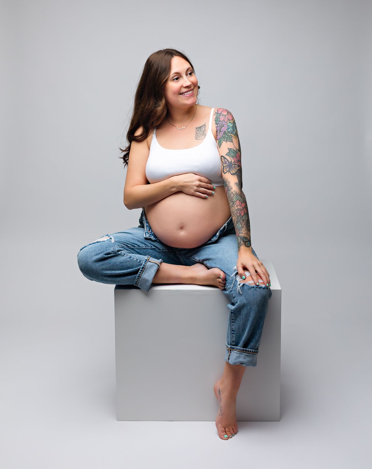 Smiling tattooed pregnant woman in white crop top and ripped jeans sitting on white cube.