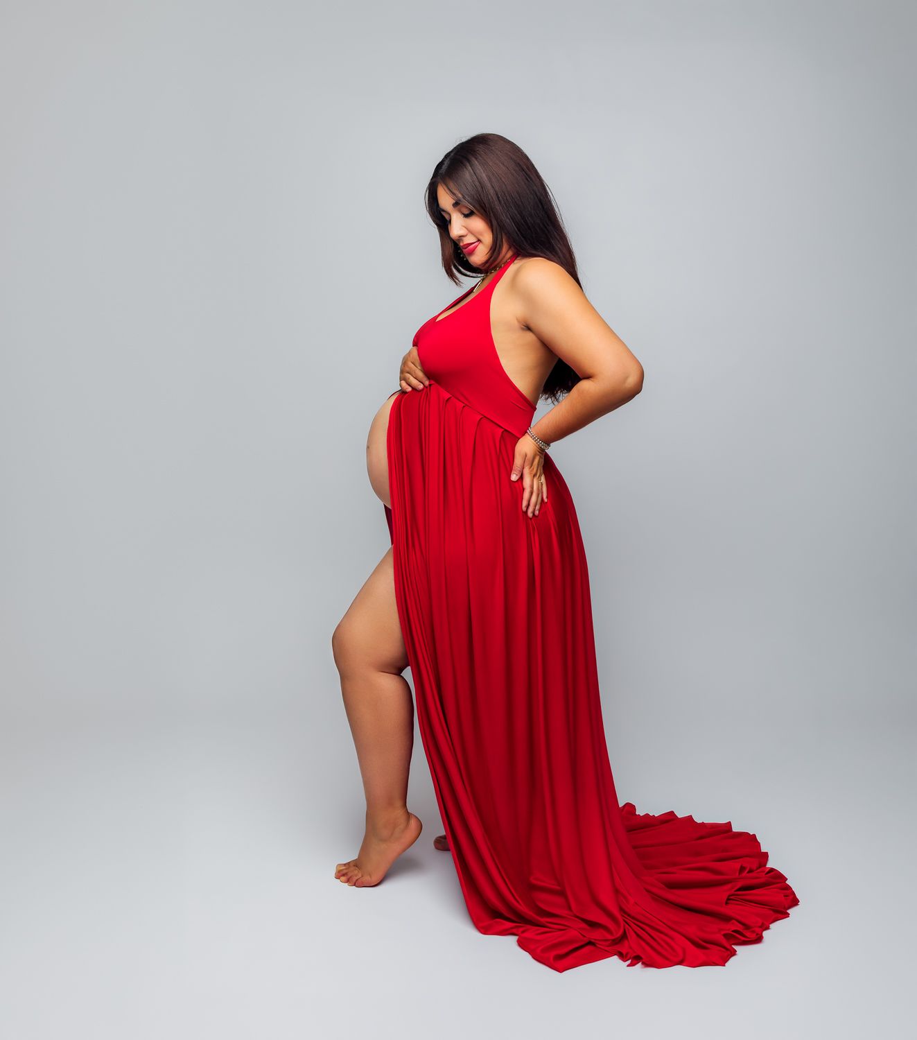 Pregnant woman in elegant red maternity gown with slit, posing sideways on gray background.