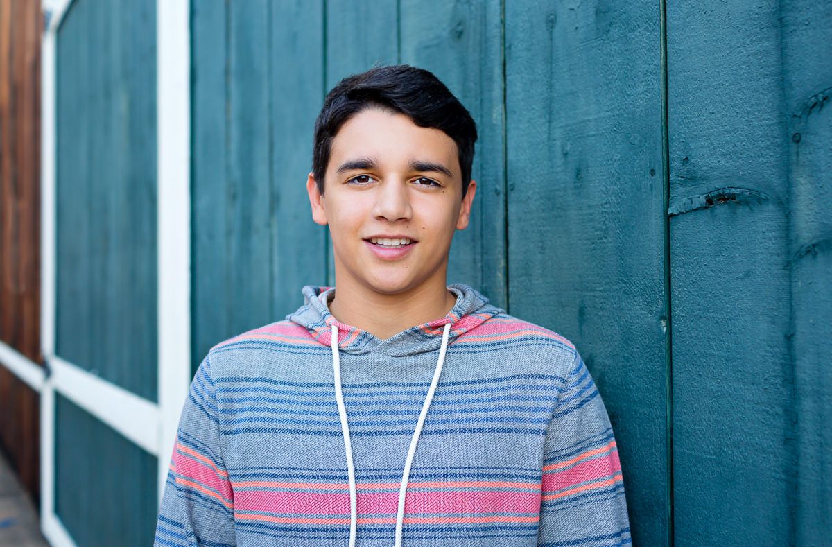 Smiling teenage boy in striped hoodie posing against teal wooden wall outdoors.