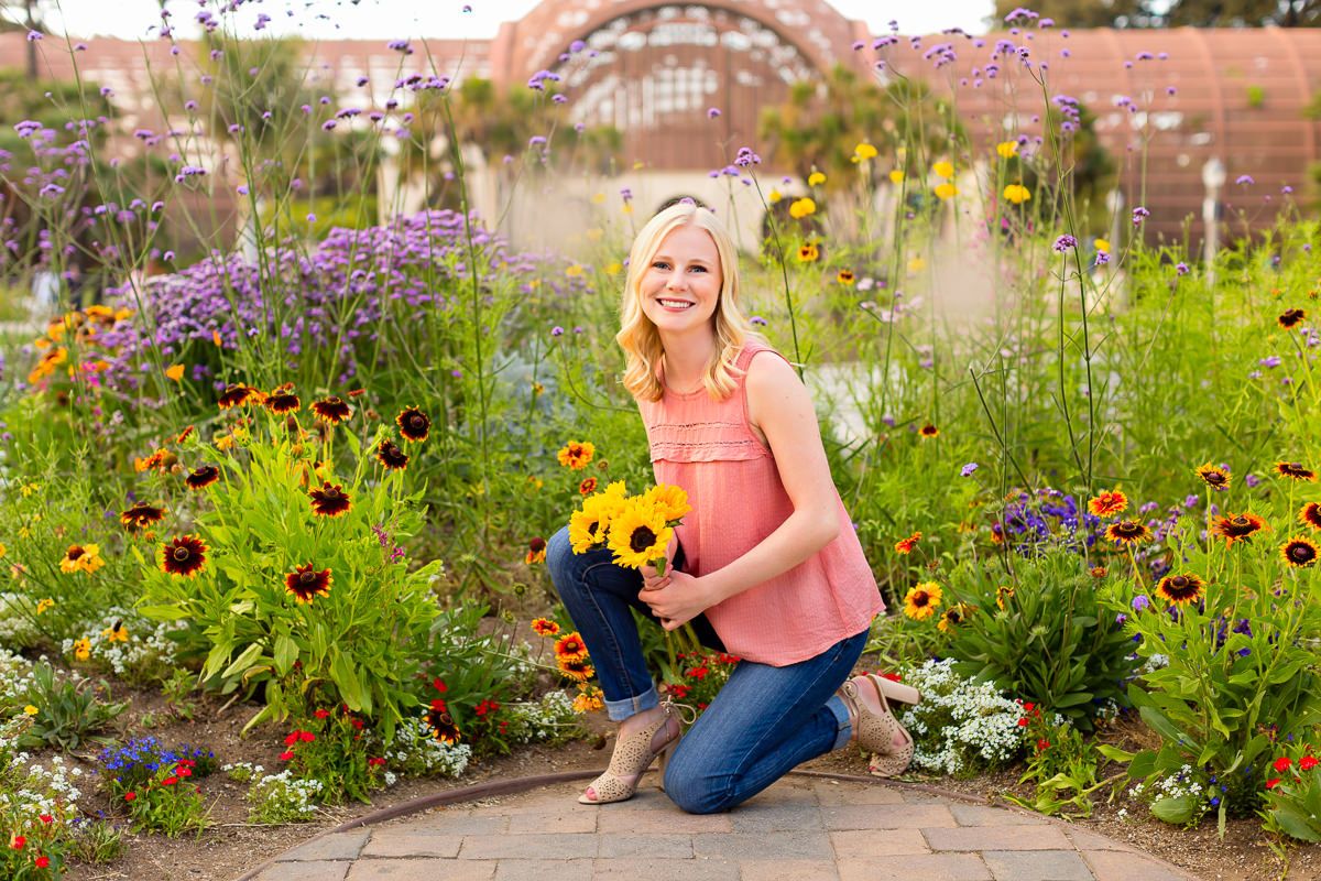 Smiling blonde woman kneeling in colorful garden holding yellow sunflowers, surrounded by vibrant blooms on brick path.