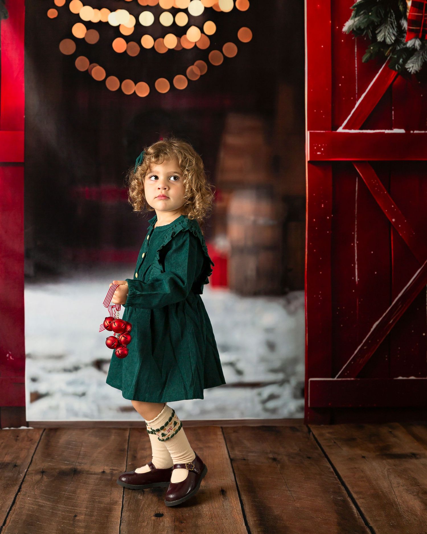 A young girl in a green dress holds red ornaments, standing in a festive barn doorway with snow and holiday lights.