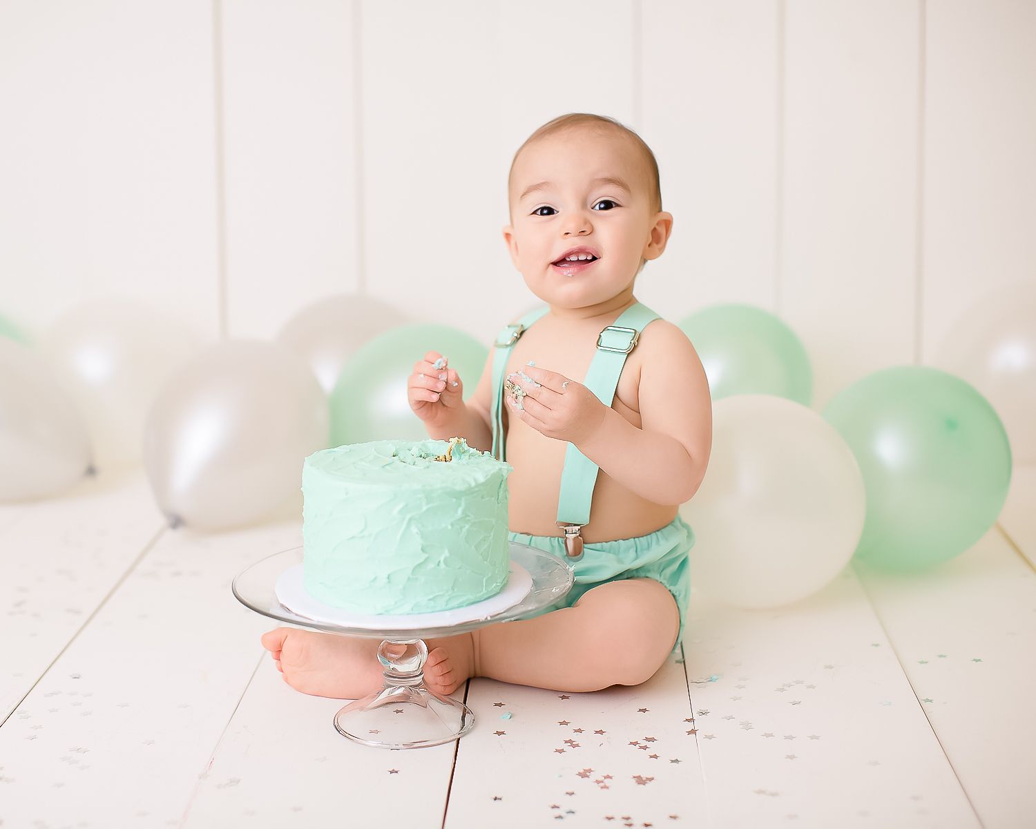 Baby in mint romper smashes first birthday cake surrounded by green and white balloons on white floor.